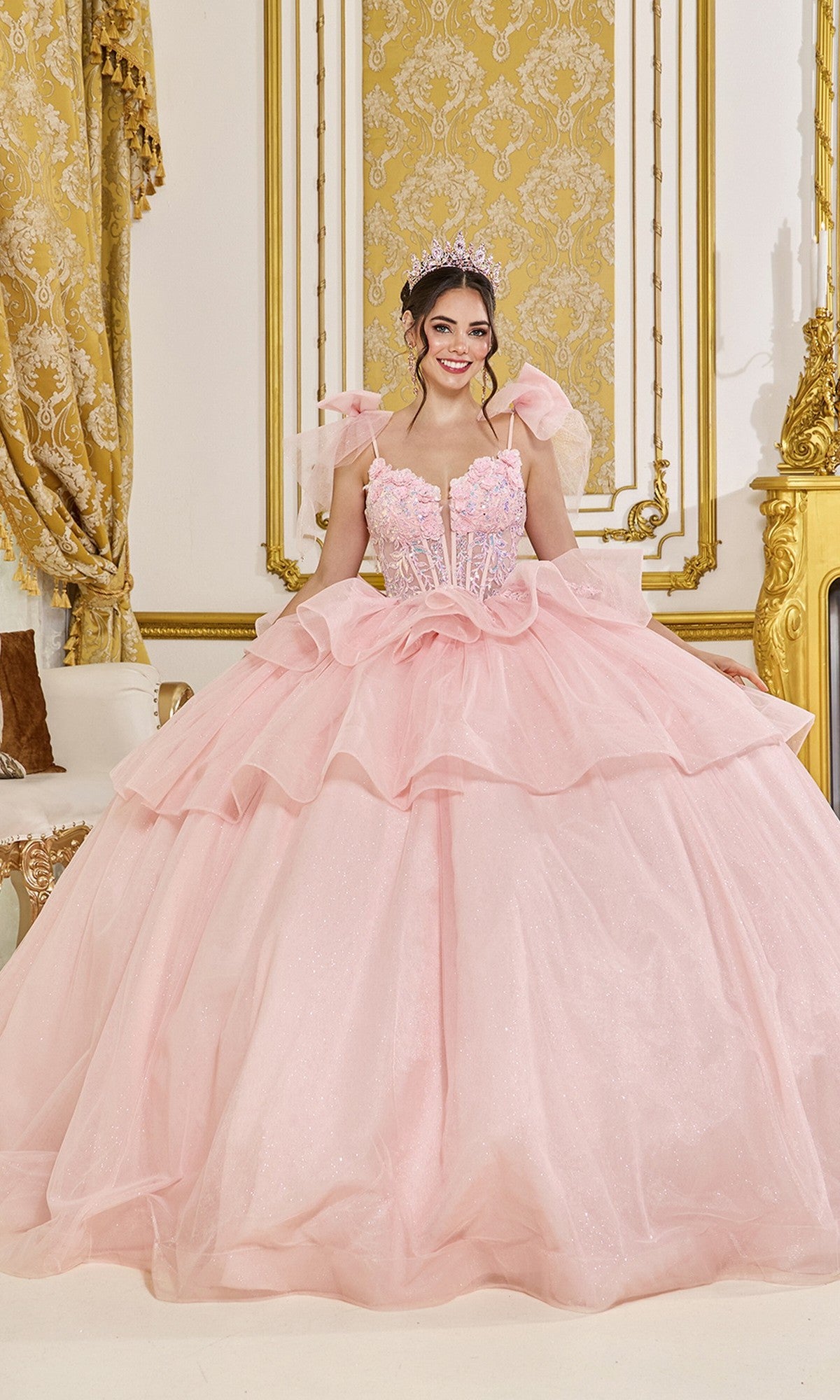 A woman poses indoors in the Quinceanera Dress 5079 by Dancing Queen in blush, featuring shoulder strap bows and a sparkling tiara, set against ornate gold and white decor.